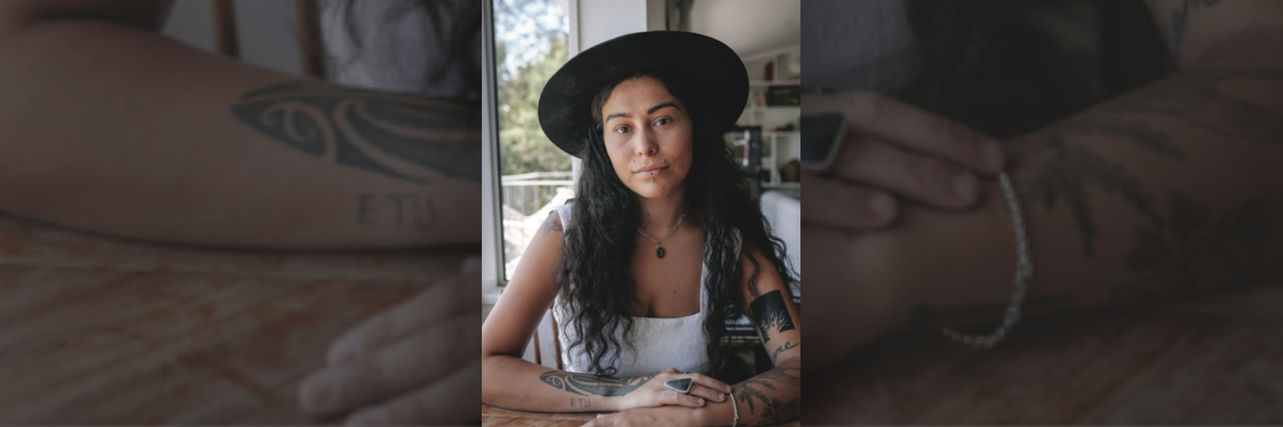 isla huia, writer, wearing a black hat sitting at a table, with crossed arms