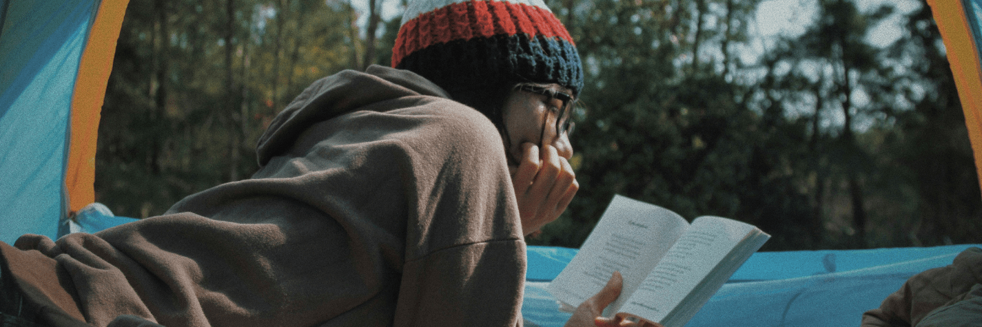 Person sitting in tent, reading. Wearing multicoloured beanie and brown hoodie.